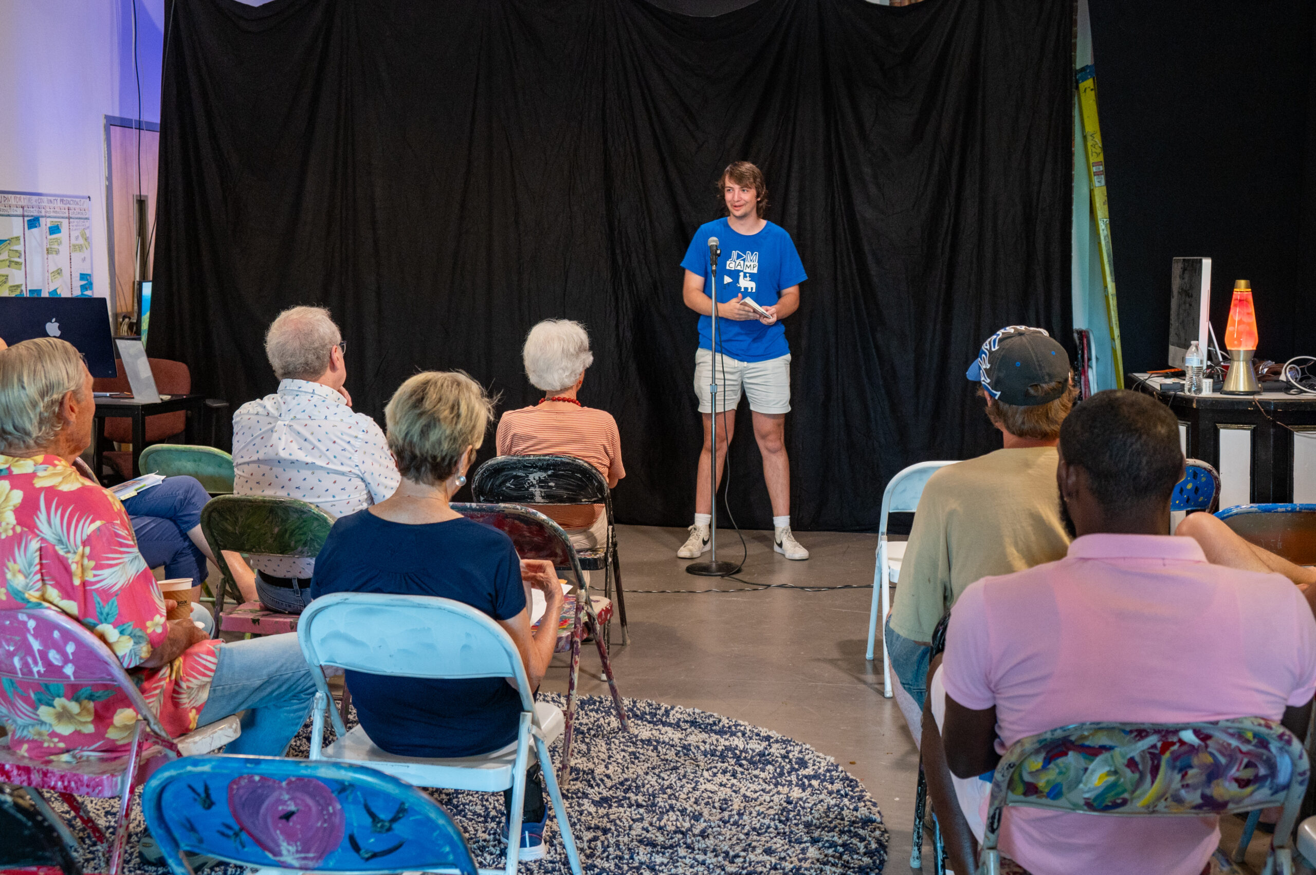 A young man with blonde hair wearing a blue teeshirt and kahki shorts standing in front of a small crowd of people giving a presentation