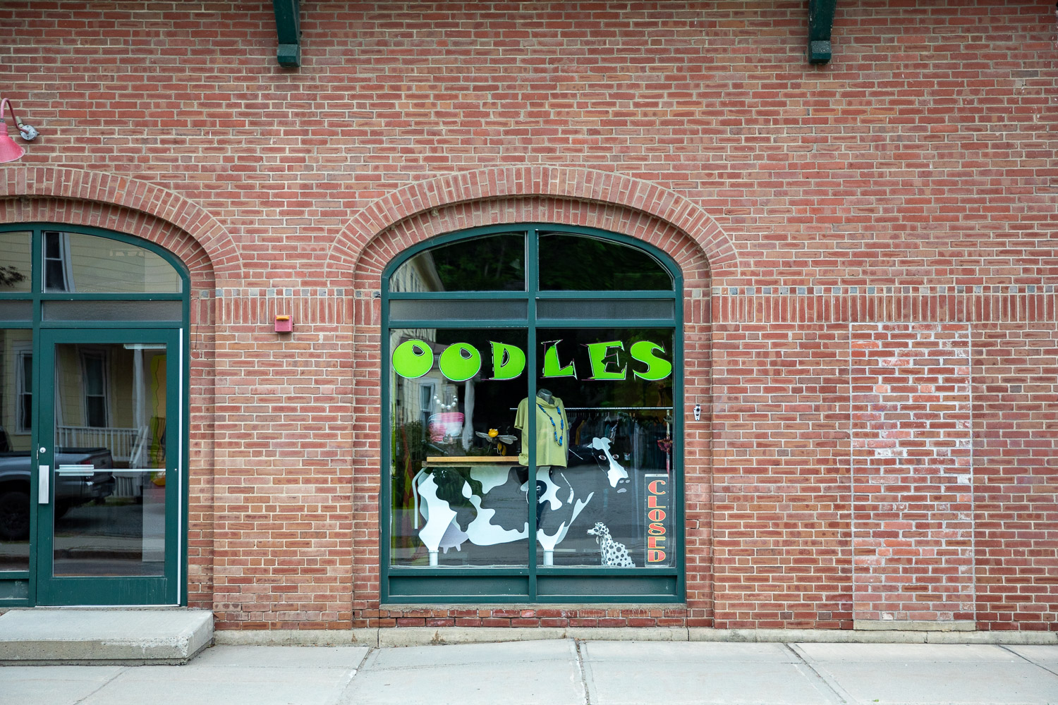 005A7503-2 A brick storefront with a large arched window displaying the name "Oodles" in bright green letters. A cow graphic decorates the lower part of the window, and a "CLOSED" sign hangs vertically on the right.