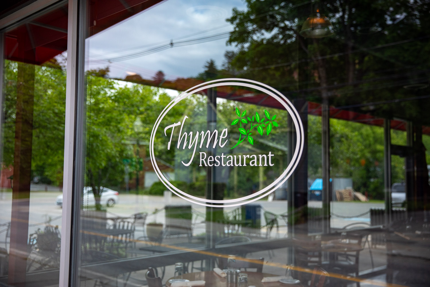 A glass window with the logo for Thyme Restaurant, featuring elegant white text and a green herb graphic, with reflections of outdoor seating and trees in the background.