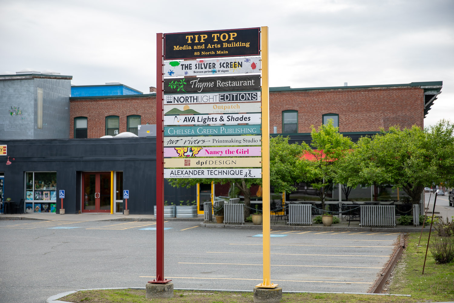 005A7512 A colorful directory sign for the Tip Top Media and Arts Building, listing various businesses including restaurants, publishers, and studios, with a red and yellow frame and buildings visible in the background.