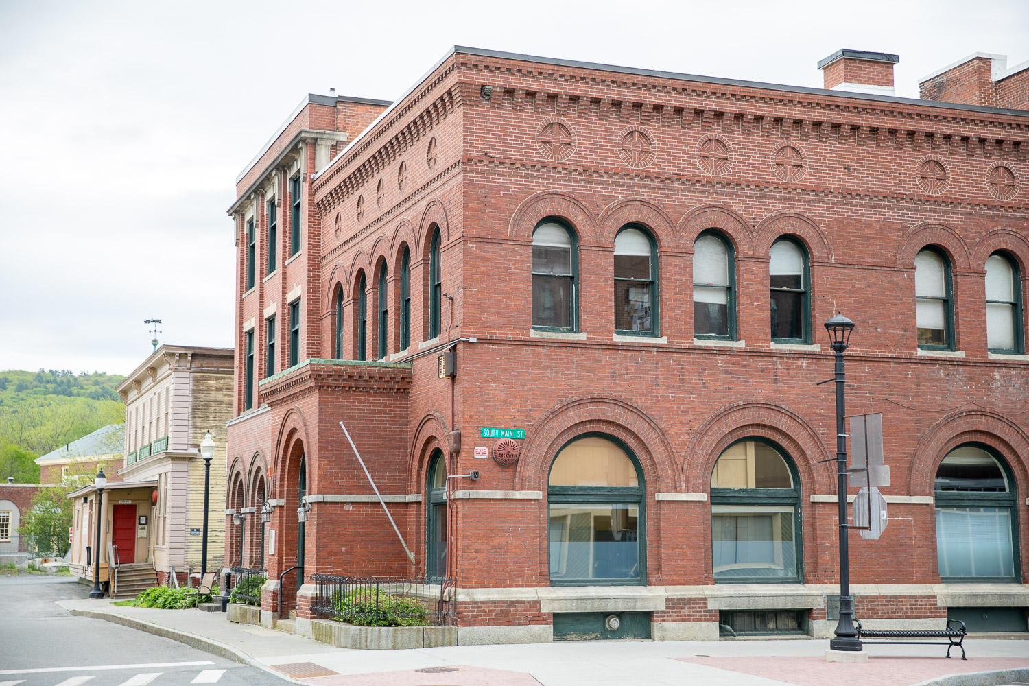 005A7522 A historic red brick building with arched windows sits on the corner of South Main Street in a small town. The architecture features decorative brickwork and green window trim. A streetlamp, bench, and sidewalk are visible in the foreground, with more buildings and green hills in the background.