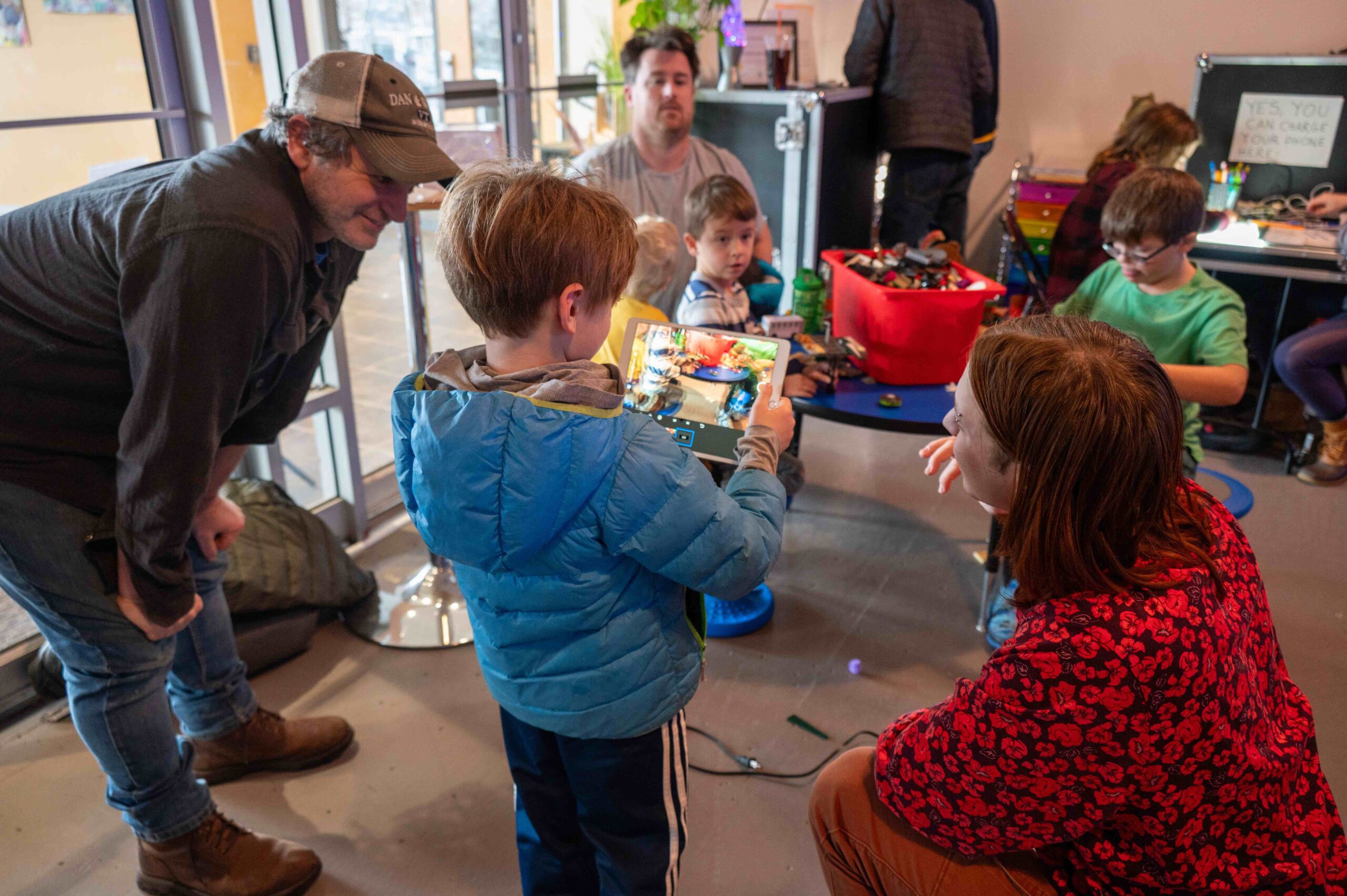 JAM Animation Pop Up Workshop - Samantha Davidson Green copy A young boy in a blue jacket holds a tablet, showing an animation he's working on to a group of adults and children gathered around him. They are in a creative workshop space with craft supplies, toys, and electronic parts on tables. Adults watch attentively and engage with the kids, who are busy building or creating.