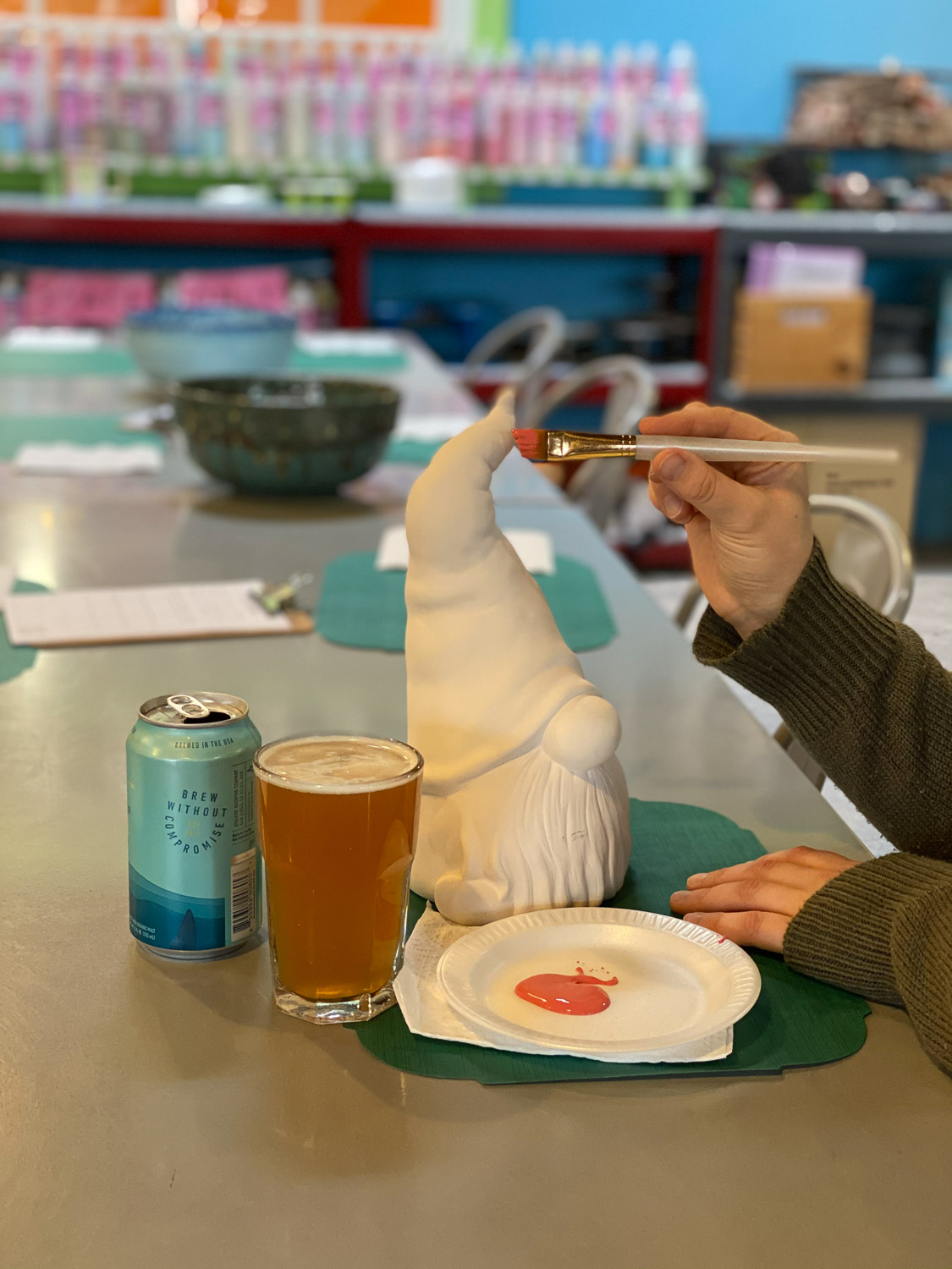 Painting with beer - Tyler Ring (Tip Top Pottery) A close up of a ceramic gnome sitting on a table. Next to the gnome there is a hand holding a paintbrush about to begin painting the gnome.